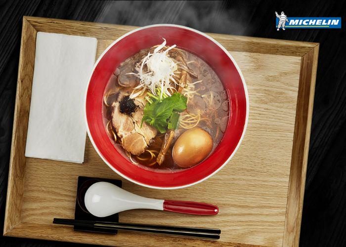 A red bowl of ramen on a wooden tray, with the word "Michelin" in the upper right corner of the image, referring to the Michelin-starred status of Tsuta, a ramen restaurant in Tokyo