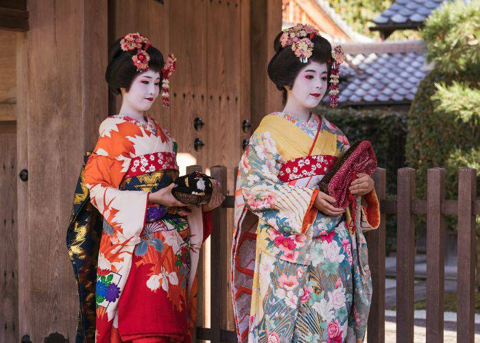 Two geisha in kimono, faces painted white, lips crimson red, dressed in traditional kimono.