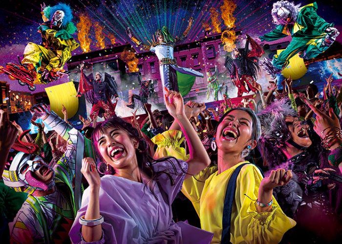 Two women dancing among a crowd of zombies, monsters, clowns, and brightly lit costumed performers onstage