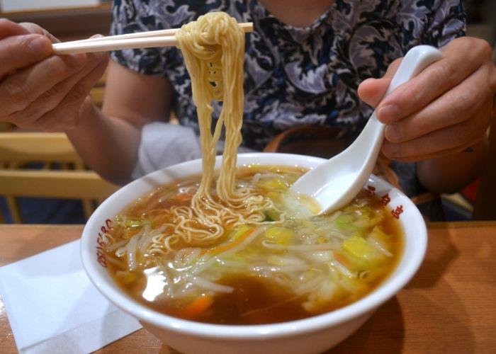 A bowl of ramen with thin straight noodles, clear broth, and lots of stir-fried veggies. Noodles being pulled up and out of the bowl by chopsticks.