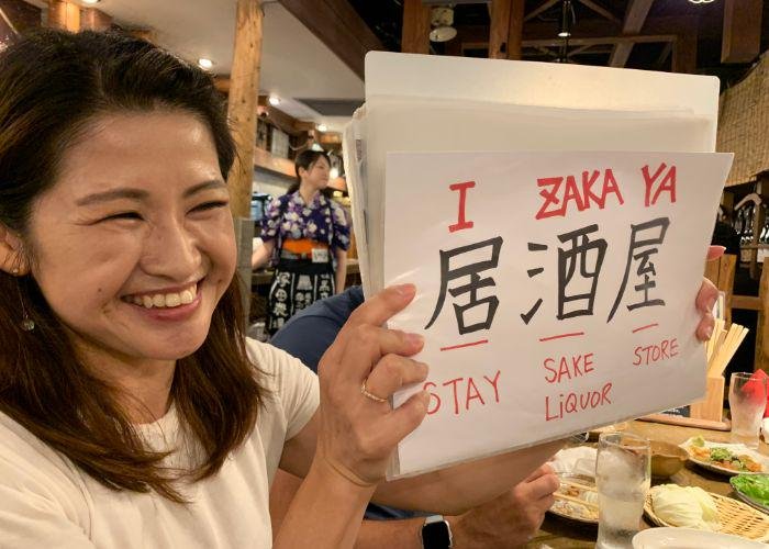 A Japanaese woman holds up a sign with the word "izakaya" written in Japanese and romaji, explaining the meaning of the word