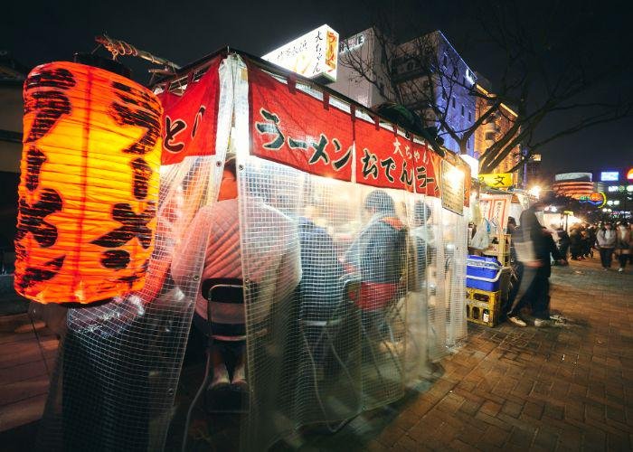 Red glowing lantern with the word "ramen" written on it in Japanese katakana, marking this Fukuoka yatai street food stall. The backs of people enjoying their ramen are visible from under the plastic tarp covering the street food stand.