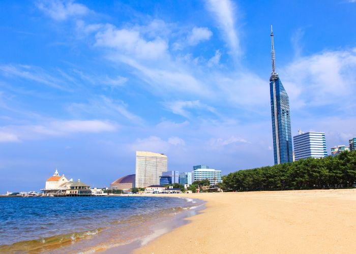 The vibrant blue waters of Fukuoka Bay meet the sandy beach, with a skyline of tall modern buildings against a blue sky and white wispy clouds