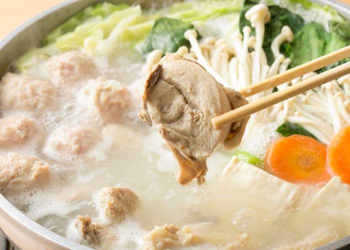 Mizutaki, a Fukuoka hot pot specialty. Pieces of chicken, carrot, mushrooms, and green veggies swim in a chicken broth. Two chopsticks hold up a piece of cooked chicken