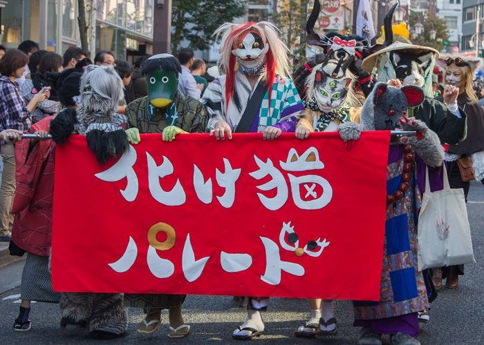 People dressed as cats, wearing cat masks and kimonos, and fuzzy cat costumes, carry a sign for Kagurazaka Bakeneko Festival