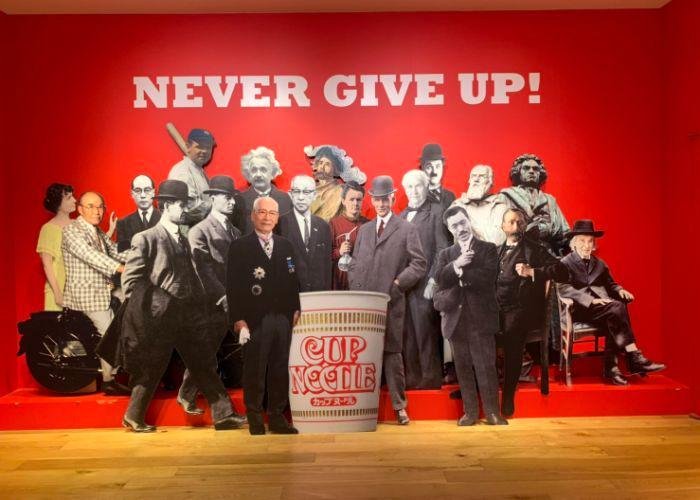 Cup Noodles Museum Interior, with a life-size cutout of Momofuku Ando, the creator of Nissin's Cup Noodle, and many other famous inventors and historical figures, with the phrase "Never Give Up!" written on the wall