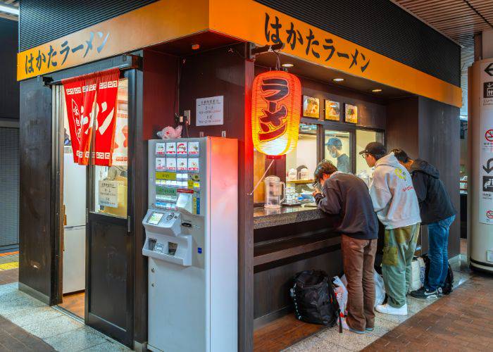 Three men standing at a ramen shop, eating ramen.