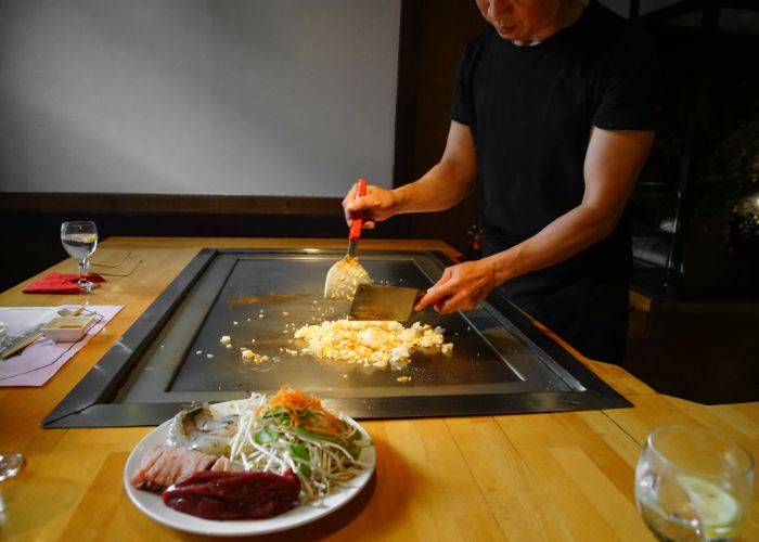 A chef using two metal spatulas to move food around a teppanyaki grill.