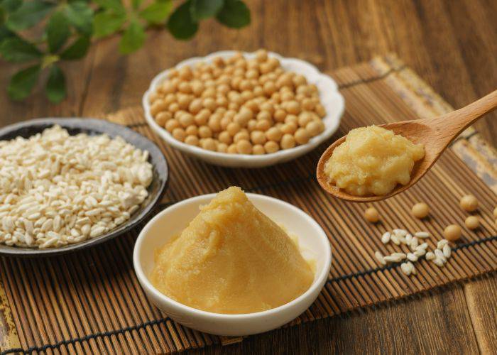 A bowl of white miso paste, including some on a wooden spoon and a bowl of the raw ingredients behind it.