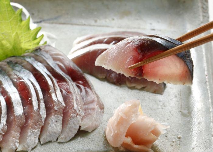 A neatly lined up row of saba (mackerel) sashimi slices with a silver skin, placed on a plate. A hand holding chopsticks picks up one piece of the sashimi
