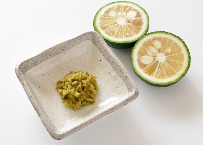 Yuzu kosho, a Japanese condiment that is made of the citrus fruit, yuzu. It is a mashed, green condiment on a plate, next to two halves of a sliced yuzu