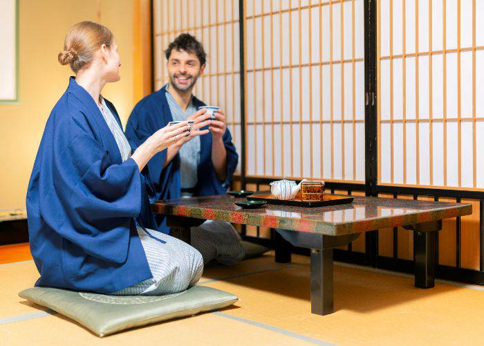 A couple smiling at each other while drinking matcha, resting on the floor of a traditional tatami mat room.