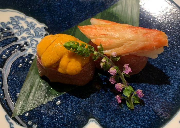 Two nigiri sushi pieces, wrapped in wagyu beef and topped with sea urchin and crab, delicately garnished with a shiso flower.