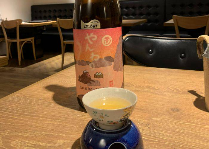 A large bottle of sake towers behind a small cup, filled with a warm, golden sake.