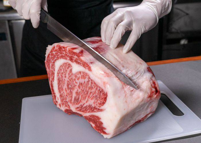 Someone in gloves slicing through a massive slab of wagyu beef with a large knife.