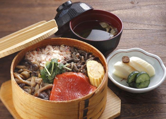 A wooden box filled with seasonal foods such as mushrooms on a table with other dishes such as soup and a dish of cucumber and bamboo shoots