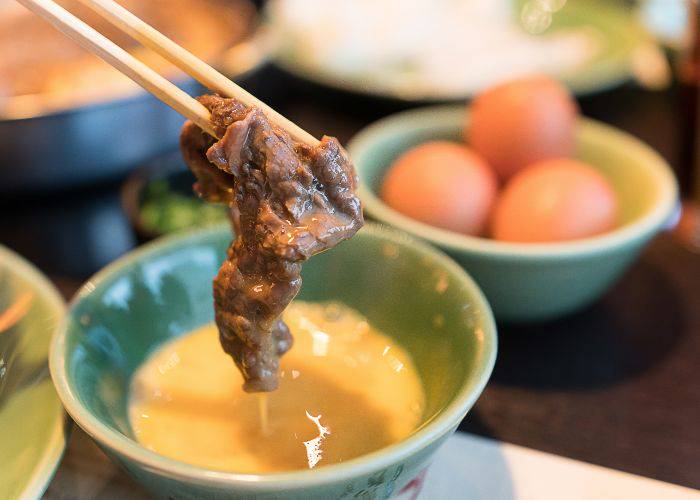 A thin slice of sukiyaki meat being dunked in raw egg before cooking.