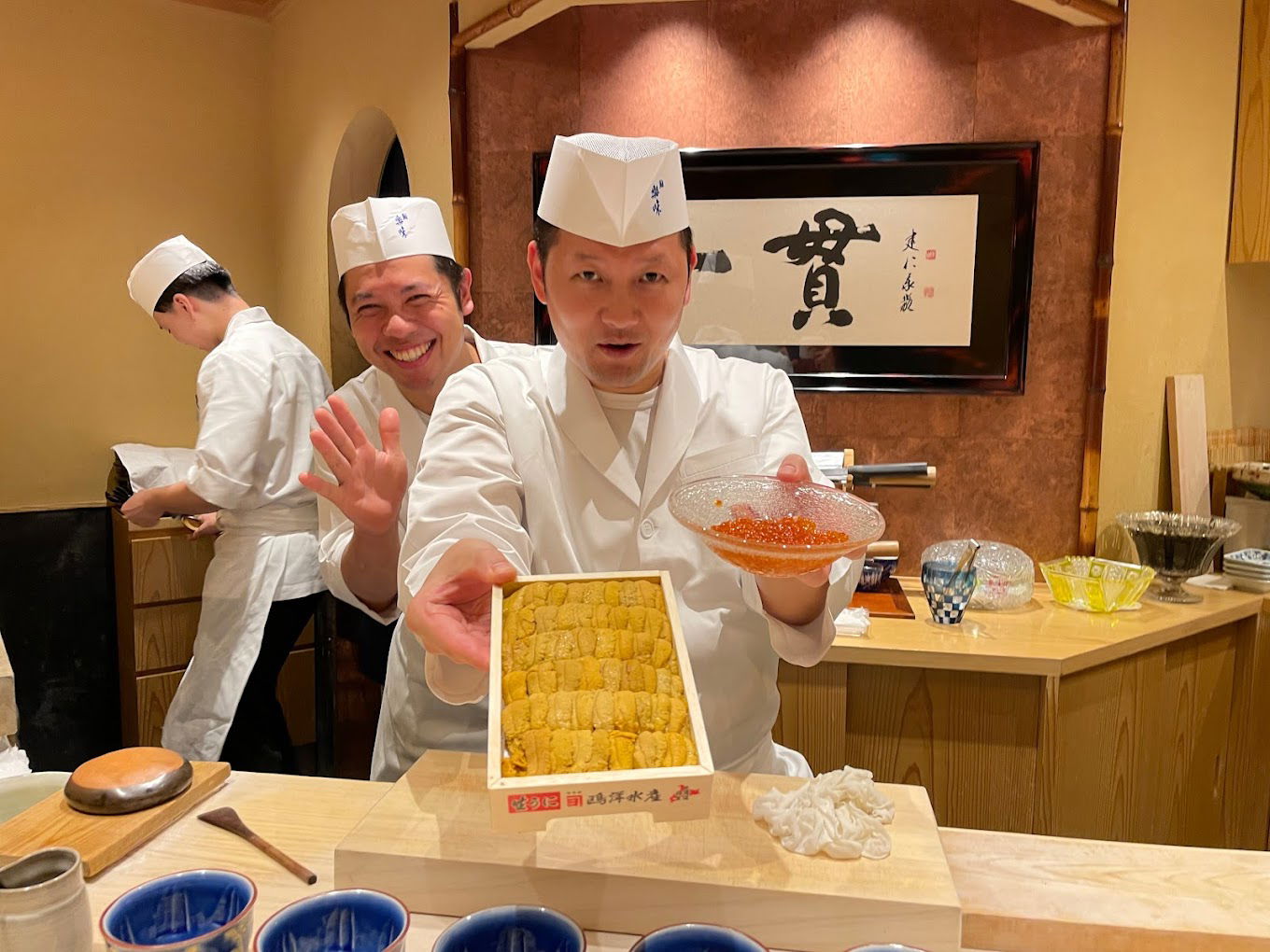 Two welcoming sushi chefs; one is offering fresh sushi while another smiling and waving.