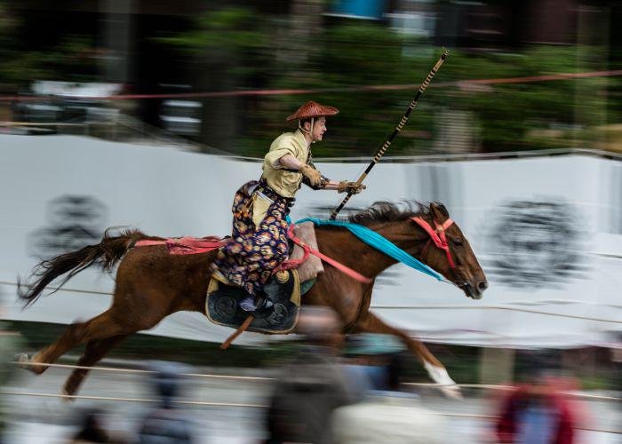 A man dressed in traditional Japanese ceremonial archery clothes riding on horseback on a brown horse 