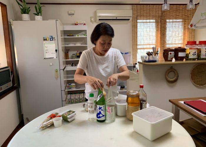 A Japanese woman stands in her kitchen behind a table laden with all manner of sake bottles, natto, miso, and other fermented foods