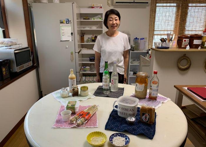 Shirley, a Japanese woman stands behind her kitchen table, smiling. There are a variety of fermented foods on the table like miso paste, soybeans, koji, sake, and natto