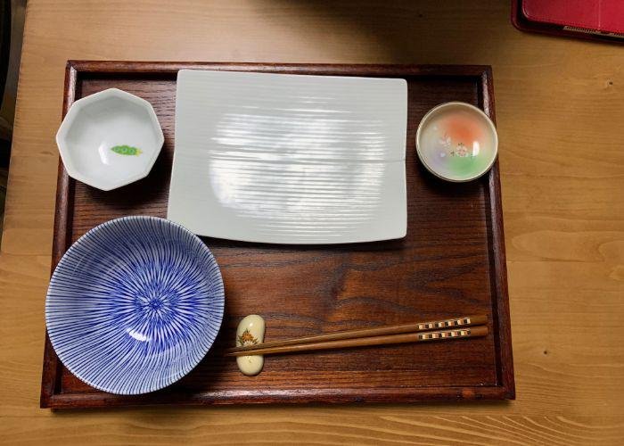 An overhead image of a wooden tray with several small dishes, a striped blue bowl, octagonal small sauce dish, chopsticks, and a rectangular white plate
