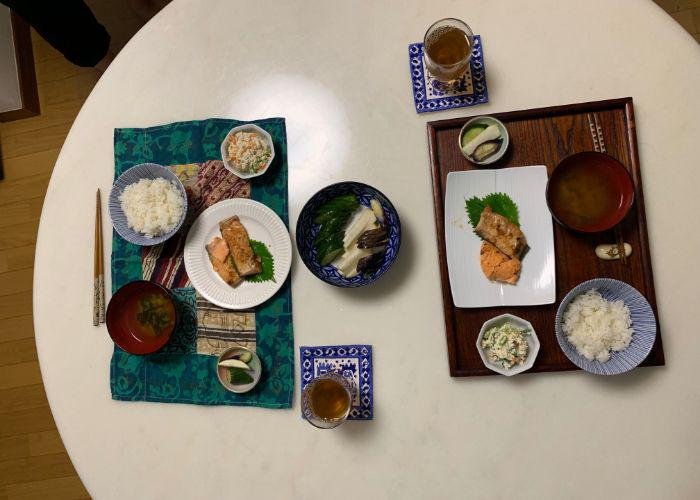 Overhead shot of a round white table with two place settings. The plates have grilled salmon, bowls of white rice, miso soup, pickled dishes, and tea