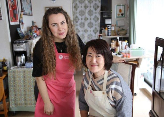 A girl with long hair in a pink apron and black shirt stands next to Miyuki Sensei, her seated cooking instructor who is also wearing a white apron in her Japanese home