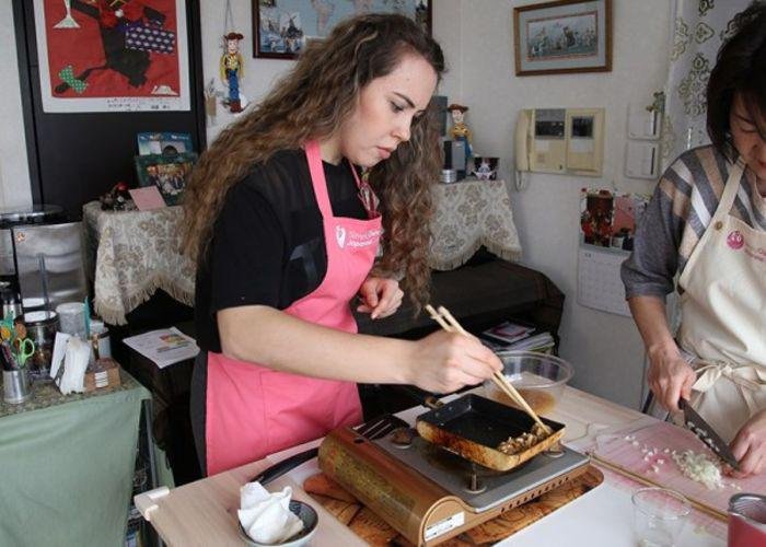 A girl in a pink apron tries to roll a tamagoyaki, Japanese omelet, as her cooking teacher looks over her shoulder