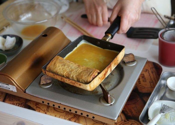 Up-close shot of a rectangular pan in which a tamagoyaki, Japanese omelette, is being rolled