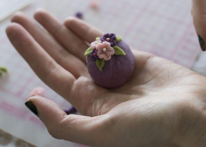 A ball of purple wagashi, decorated with cute pink flowers and green leaves, is held in the palm of a woman's hand