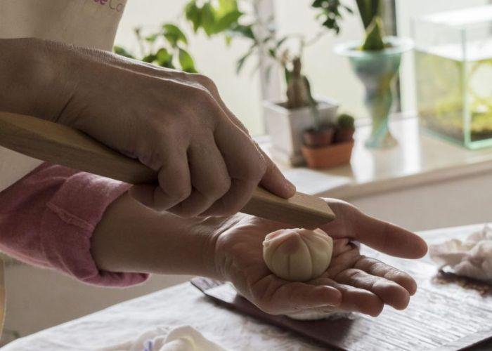 The cooking instructor holds a piece of wagashi in her hand, and uses a long wooden tool to make indents in it, creating the shape of flower petals