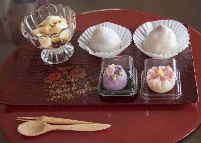 Two balls of mochi, two pices of wagashi are on a red tray with a small wooden spoon and fork.
