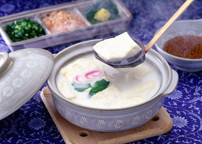A Japanese nabe (pot) is dipped into with a ladle, picking up a large piece of delicate white tofu, also known as "onsen yudofu"