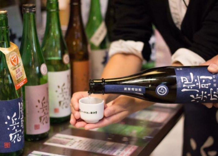 A line-up of different Japanese sake (nihonshu) from various breweries in the backround, and someone pouring into a small sake cup in the foreground