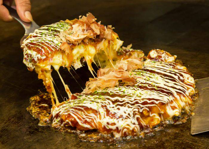 Okonomiyaki is being taken off the grill; melted cheese inside extends down to the grill.