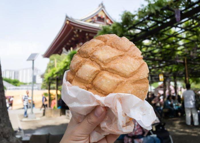 A melon pan being held up against Asakusa's iconic Senso-ji Temple.