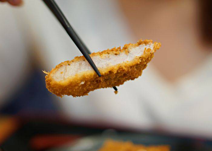 A slice of breaded tonkatsu pork cutlet, held in the air by chopsticks.