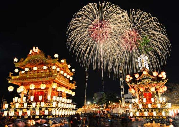 An image of two brightly decorated Japanese floats adorned in lanterns that are lit up at night with colourful fireworks exploding in the background of the night sky