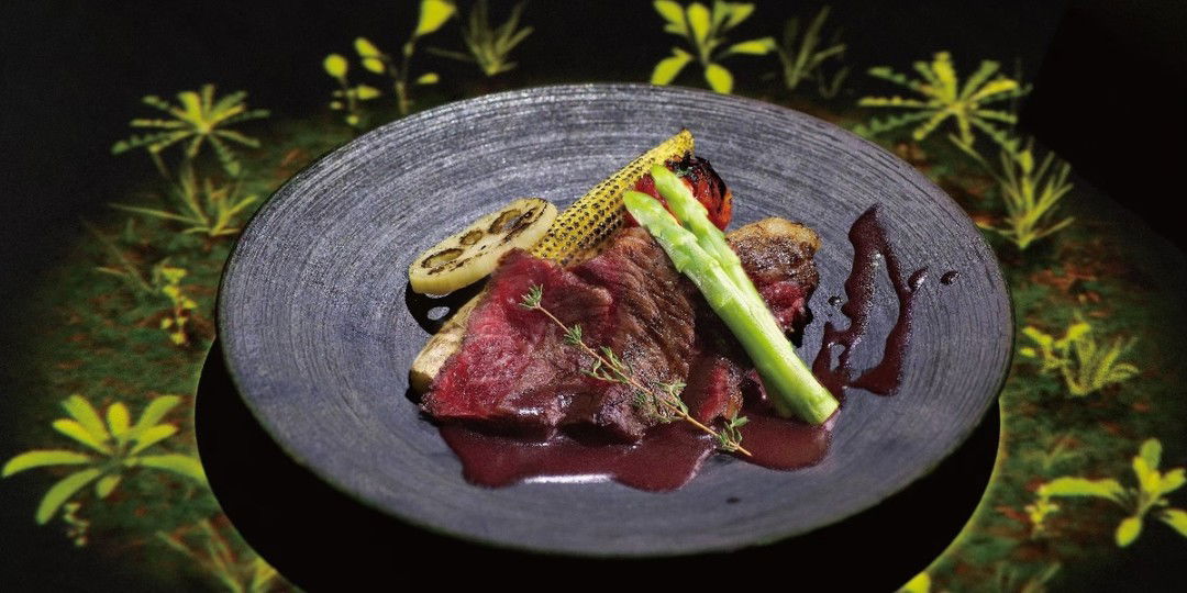 A plate of Kobe beef steak and a variety of vegetables sitting on a table that has projection mapping of flowers displayed on it at Farm to Table Kanbe