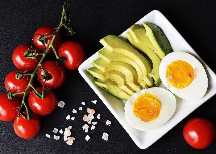 A spread of tomatoes on the vine, hard-boiled egg, and sliced avocados on a white plate