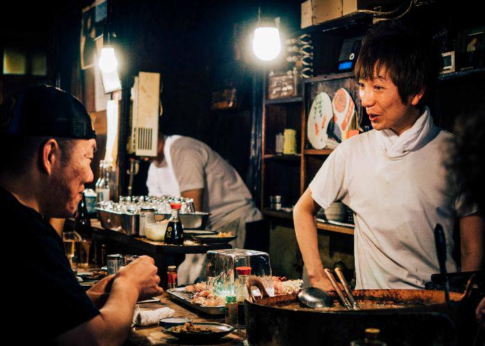 The izakaya barkeeper chats with a customer, over a table laden with grilled meat skewers