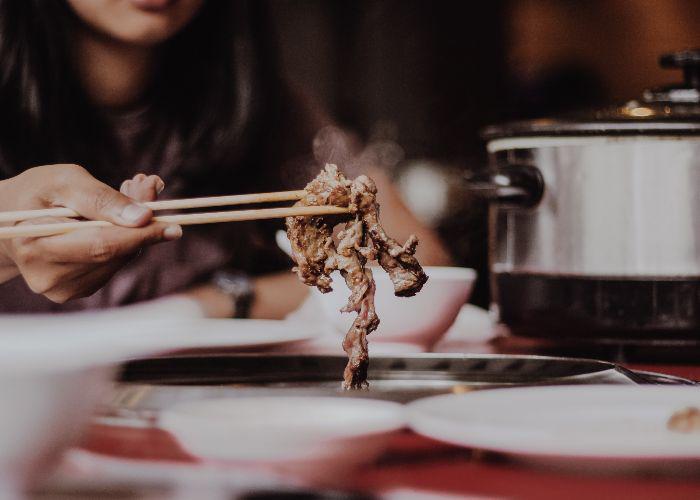 A woman holds out a piece of grilled meat, freshly cooked and steaming, between her chopsticks