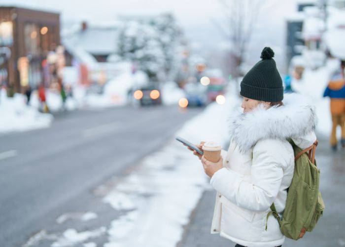 A woman in a warm jacket and hat, using her phone on the side of a snowy road in Niseko.