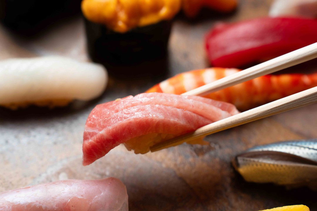 Chopsticks picking up salmon sushi — behind it, more types of sushi.