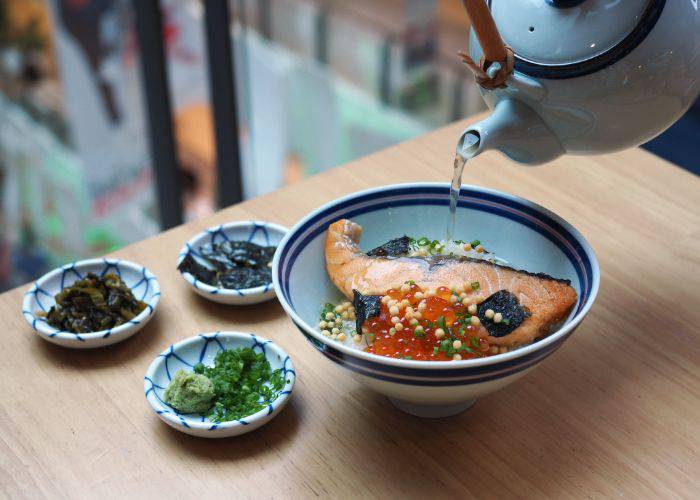 A bowl of salmon ochazuke with tea being poured on top. Three side dishes sit next to the bowl