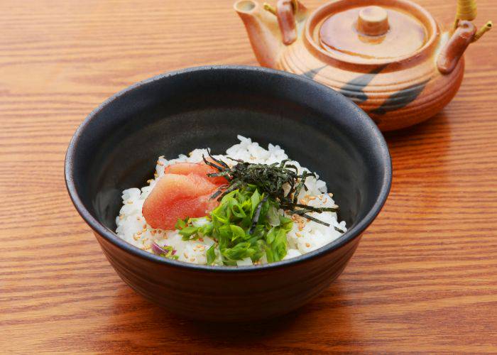 A bowl of tarako ochazuke topped with nori and other vegetables. A kettle sits behind the bowl