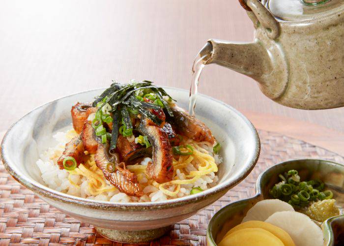 A bowl of unagi-chazuke with tea being poured from a kettle on top. It is topped with vegetables and kinshi tamago, a type of shredded egg