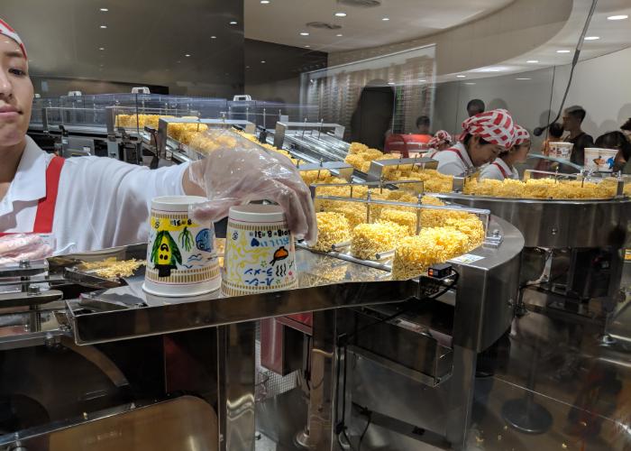 A long conveyor belt of Cup Noodles noodles comes down a conveyor belt and is packaged by Cup Noodle Museum staff