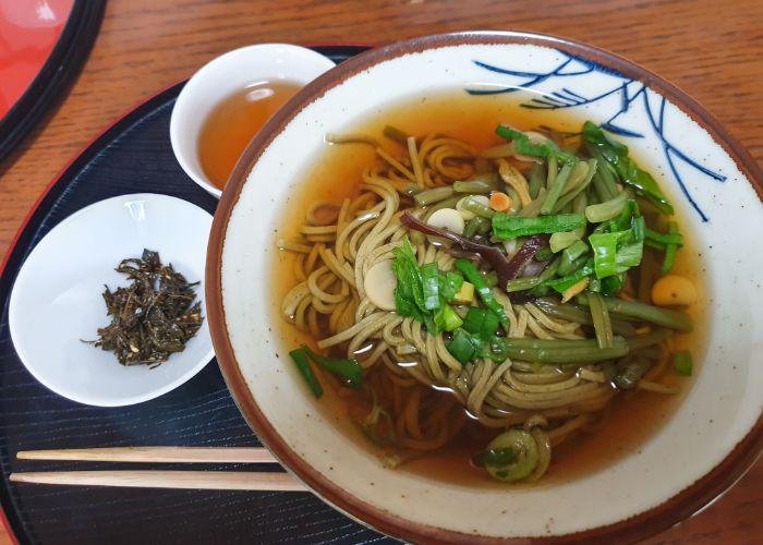 A bowl with seasonal vegetables soba with green tea leaves with sesame on the side and some green tea